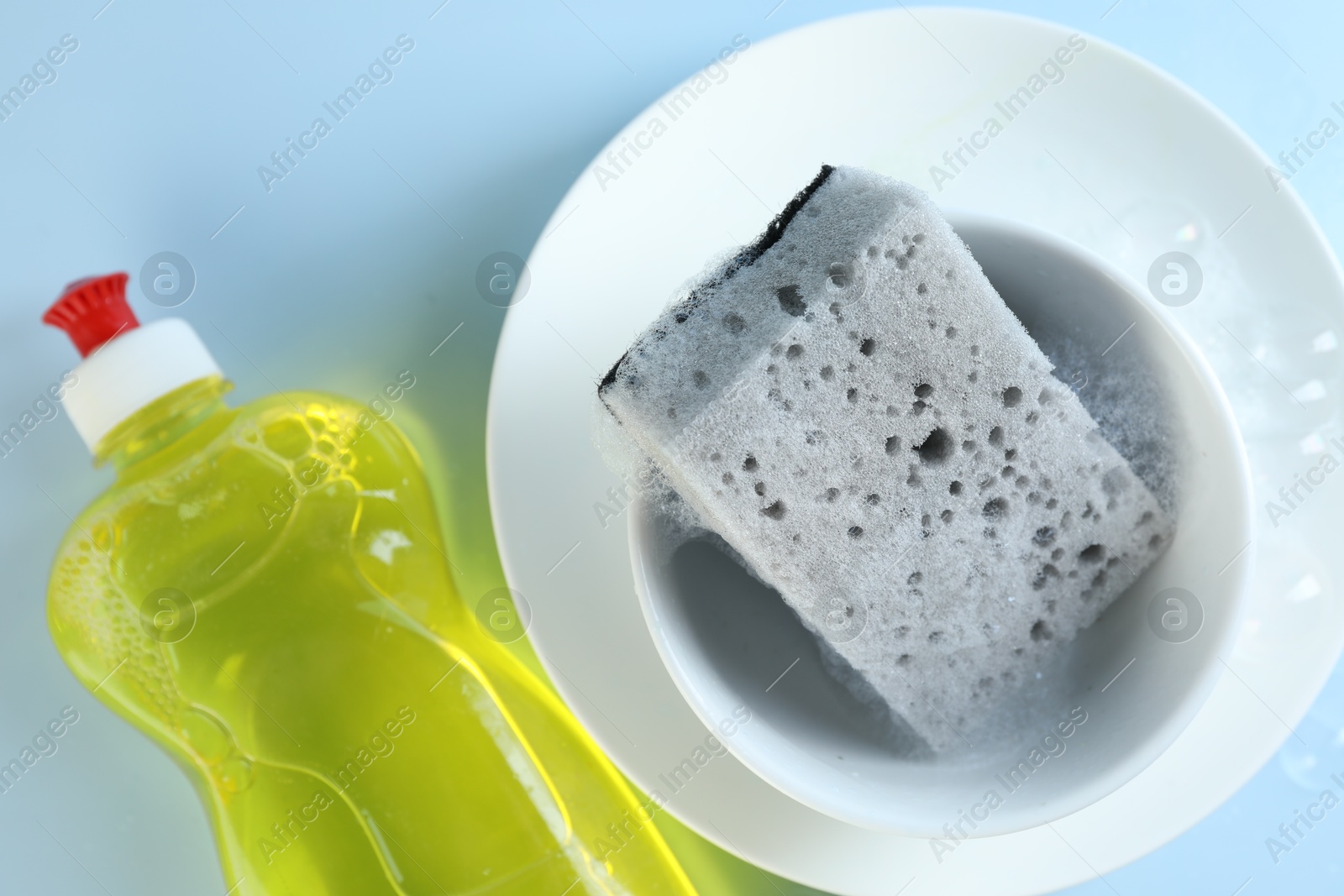 Photo of Sponge with foam, dishware and bottle of detergent on light blue background, flat lay