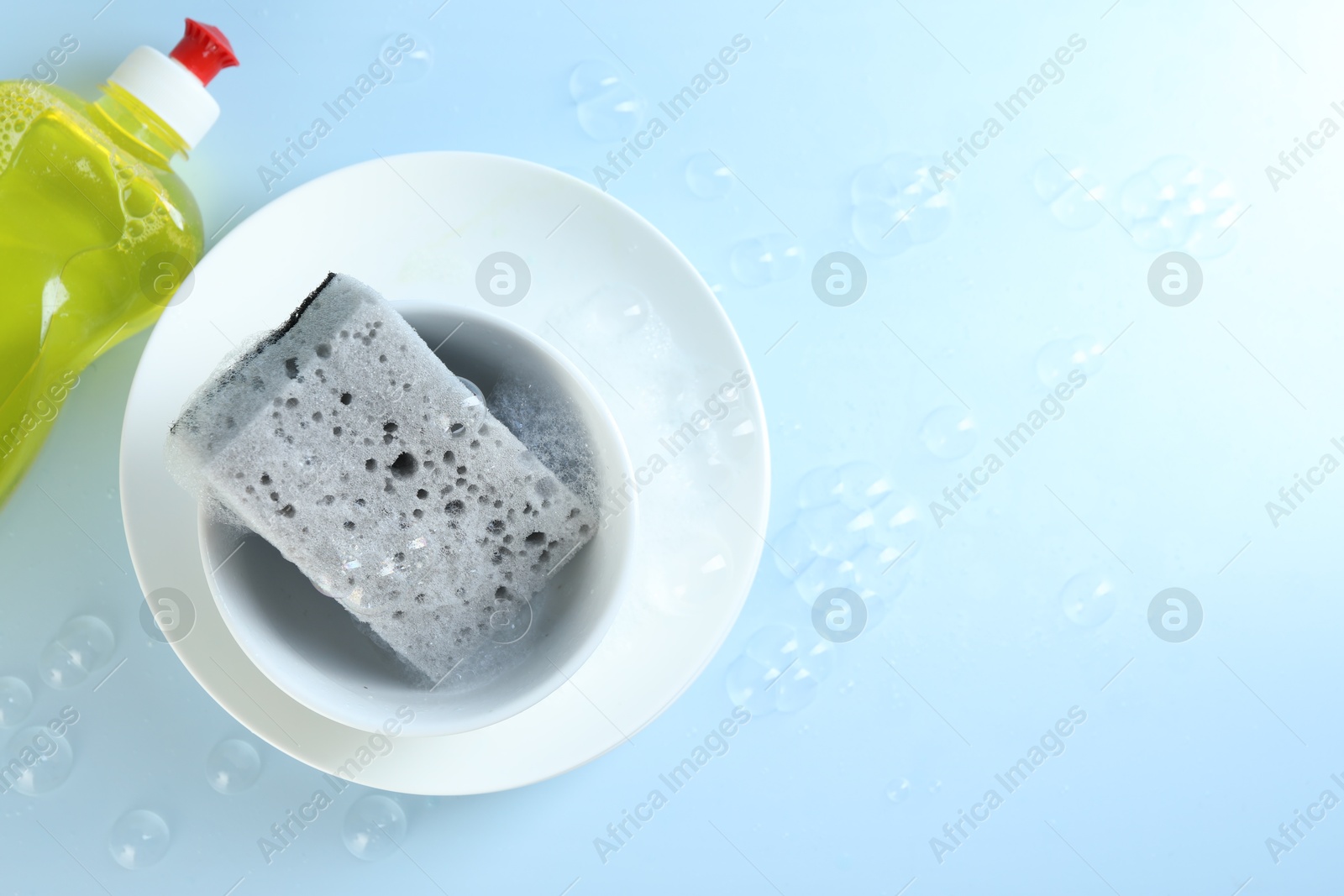 Photo of Sponge with foam, dishware and bottle of detergent among bubbles on light blue background, flat lay. Space for text