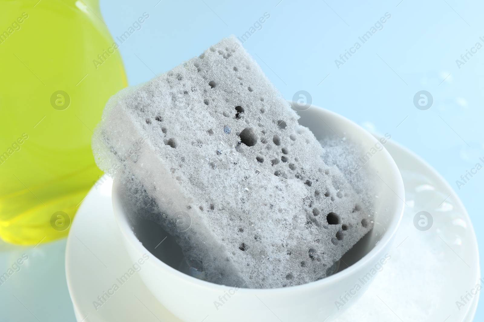 Photo of One sponge with foam and dishware on light blue background, closeup