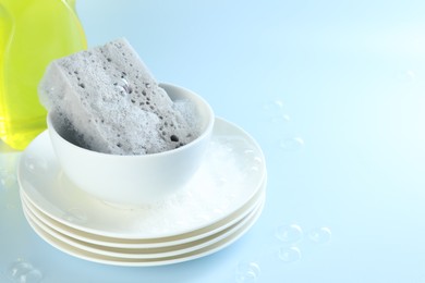 Photo of Sponge with foam, stack of dishware and bubbles on light blue background, closeup. Space for text