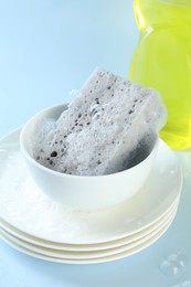 Photo of Sponge with foam, stack of dishware and bubbles on light blue background, closeup