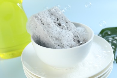 Photo of Sponge with foam and stack of dishware among flying bubbles on light blue background, closeup