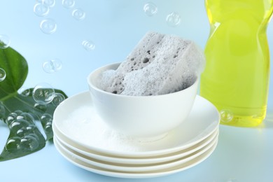 Photo of Sponge with foam and stack of dishware among flying bubbles on light blue background, closeup