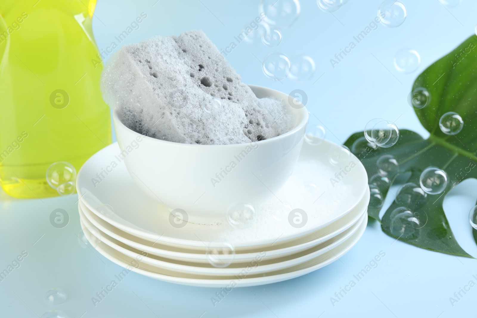 Photo of Sponge with foam and stack of dishware among flying bubbles on light blue background, closeup