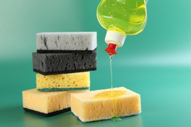 Photo of Pouring detergent from bottle onto sponge on green background, closeup