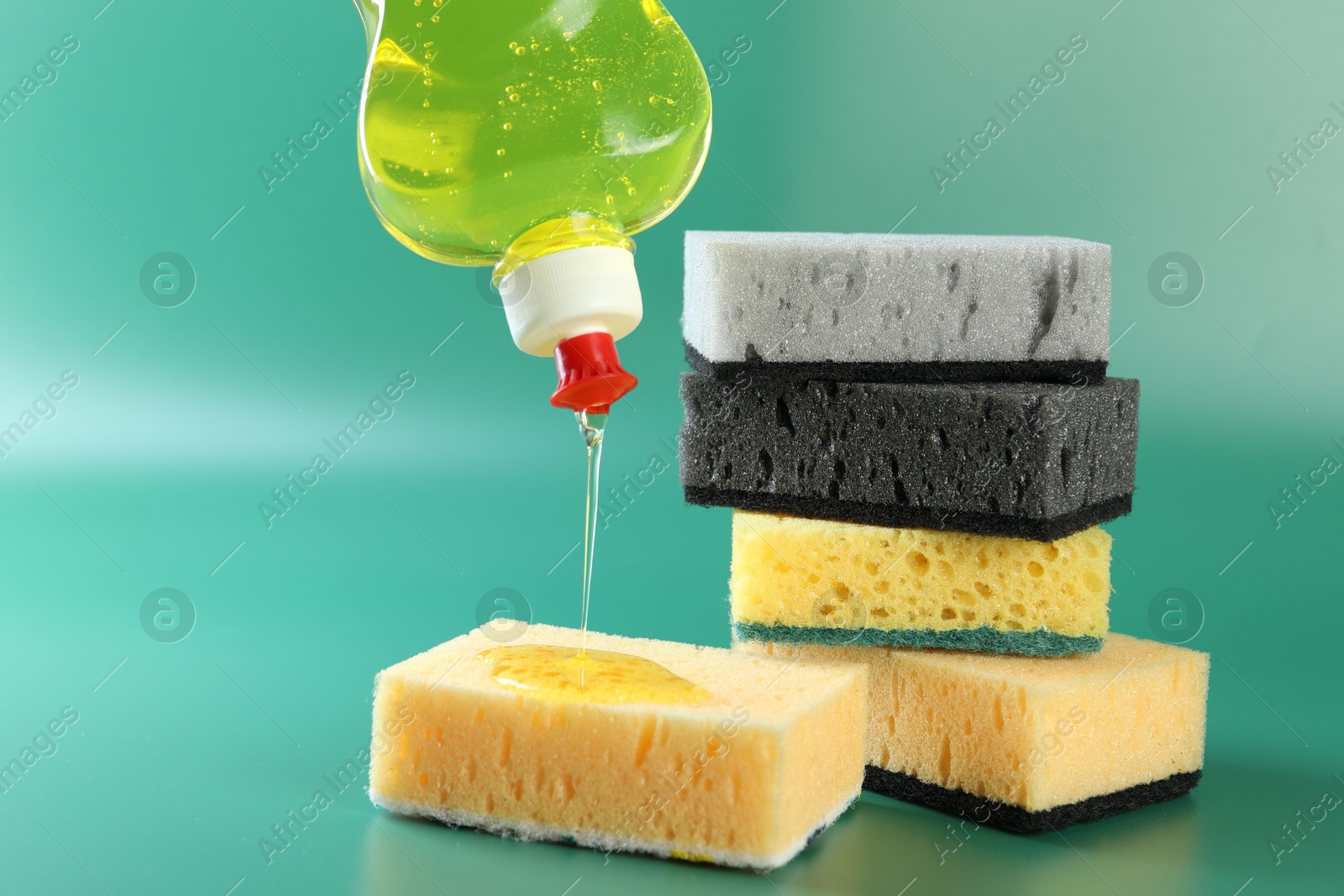 Photo of Pouring detergent from bottle onto sponge on green background, closeup