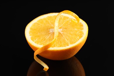 Photo of One curly orange peel and half of fruit on black mirror surface, closeup