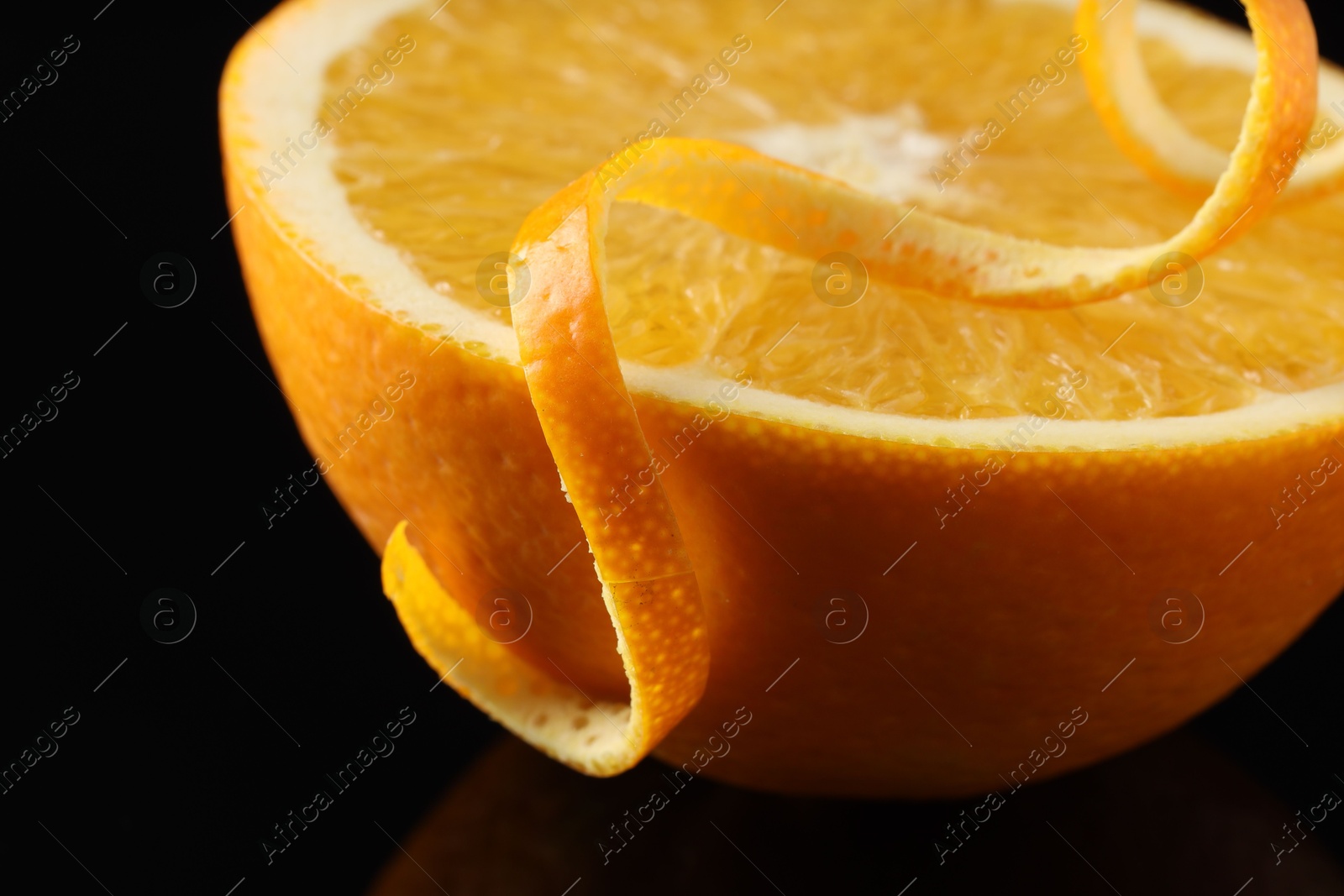 Photo of One curly orange peel and half of fruit on black mirror surface, closeup