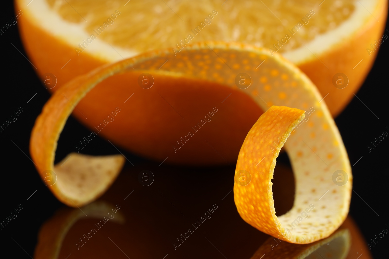 Photo of One curly orange peel and half of fruit on black mirror surface, closeup