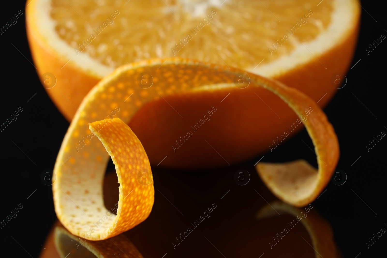 Photo of One curly orange peel and half of fruit on black mirror surface, closeup