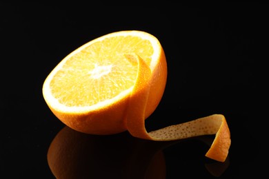 Photo of One curly orange peel and half of fruit on black mirror surface, closeup