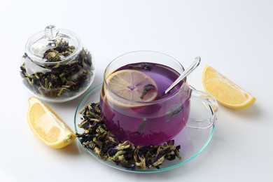 Photo of Delicious butterfly pea flower tea in glass cup, lemon and dry petals on white table , closeup