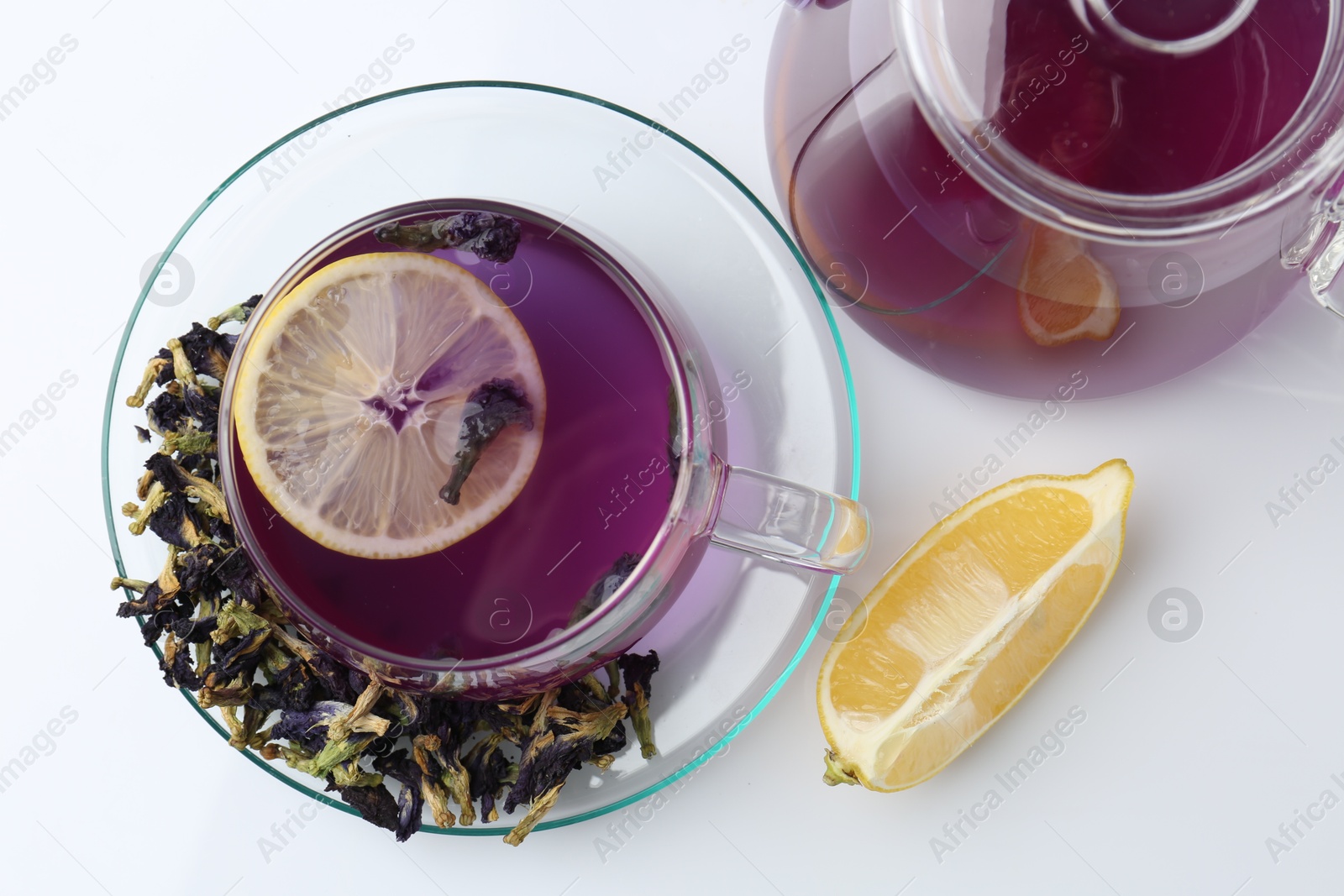 Photo of Delicious butterfly pea flower tea in glass cup, lemon, teapot and dry petals on white table , flat lay