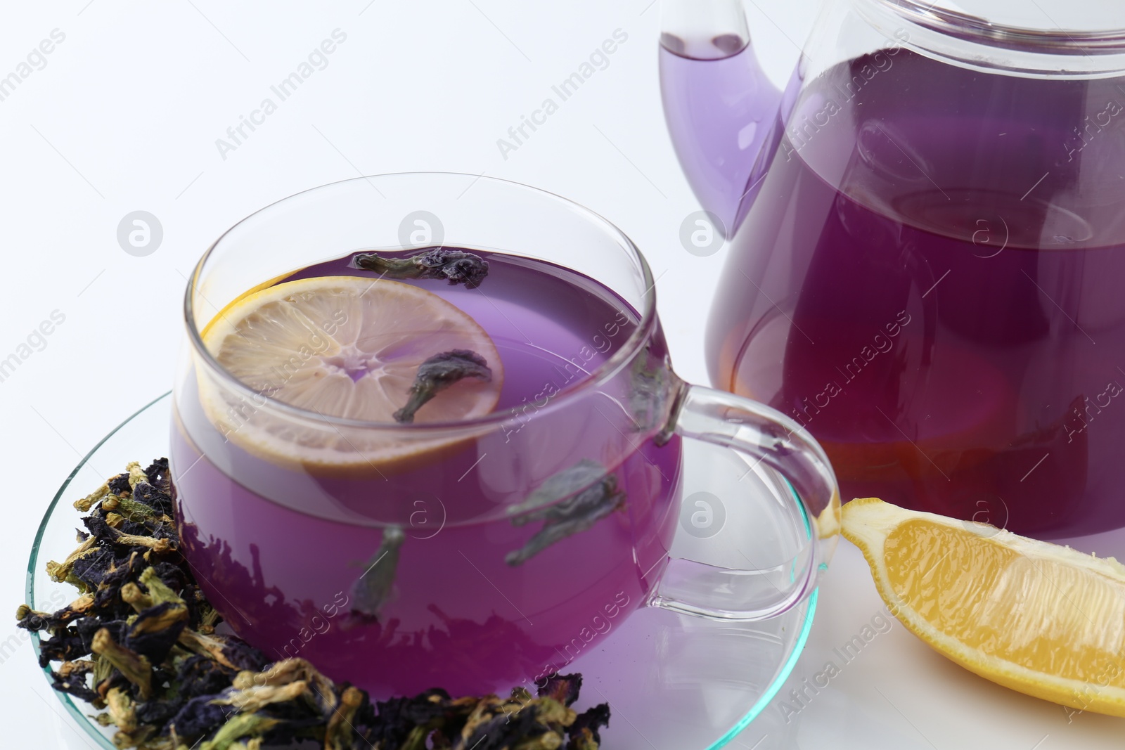 Photo of Delicious butterfly pea flower tea in glass cup, lemon, teapot and dry petals on white table, closeup