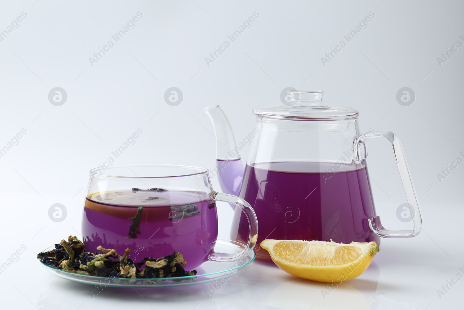 Photo of Delicious butterfly pea flower tea in glass cup, lemon, teapot and dry petals on white table