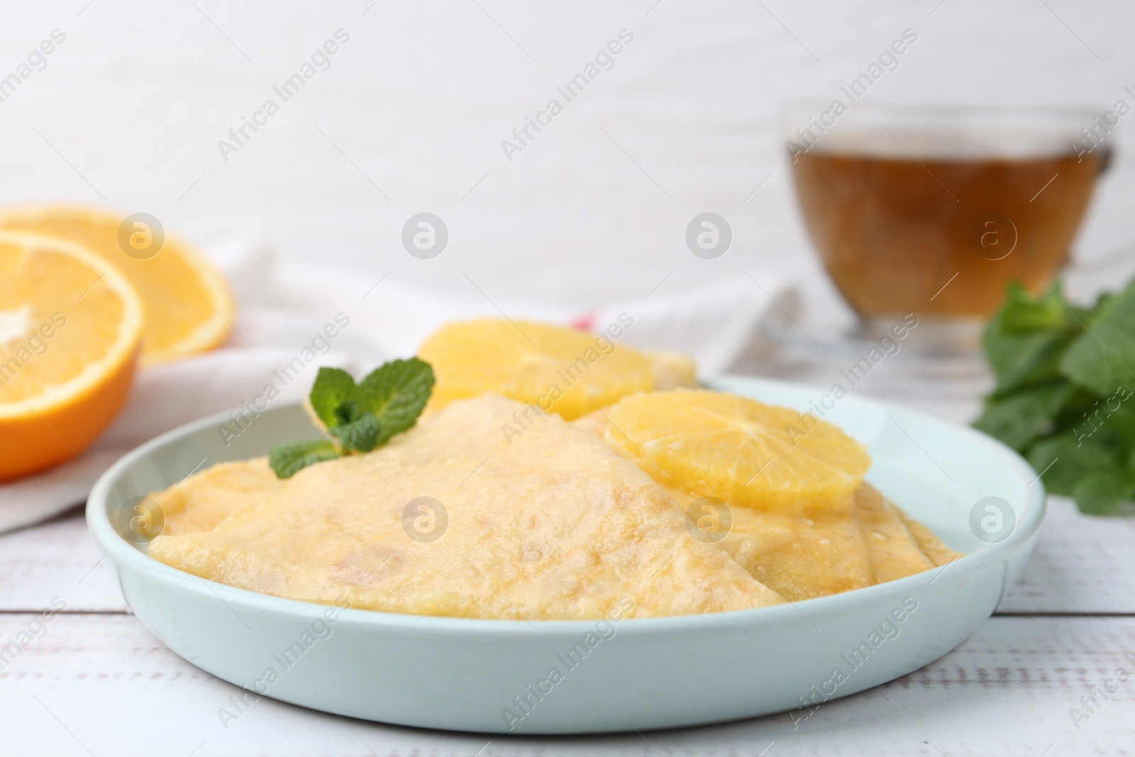 Photo of Delicious Crepes Suzette, mint, tea and oranges on white wooden table, closeup