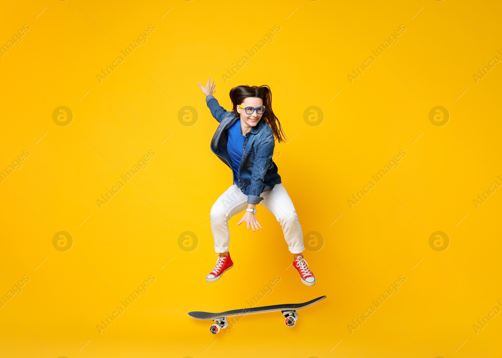 Image of Beautiful woman with skateboard in air on orange background. Performing trick