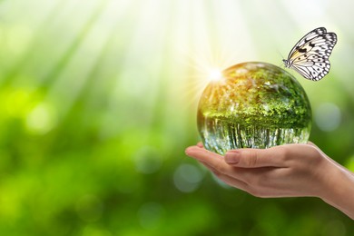 Image of Save Earth. Woman holding ball lens with overturned reflection of green forest in sunlight, closeup. Butterfly on crystal globe