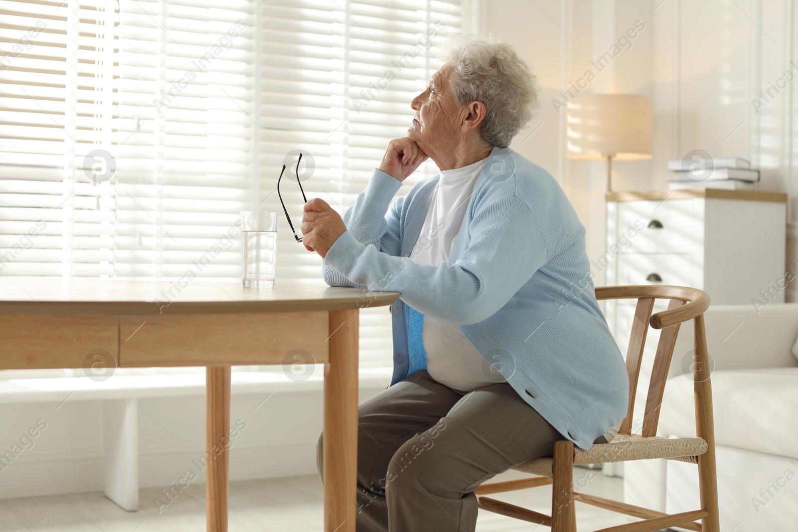 Photo of Lonely senior woman looking out window at table with glass of water indoors