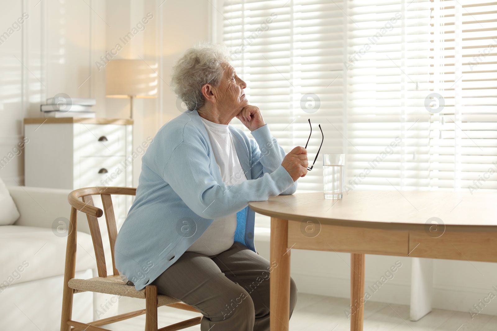 Photo of Lonely senior woman looking out window at table with glass of water indoors