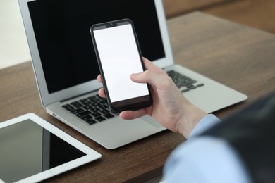 Photo of Businessman using smartphone near laptop and tablet at wooden table indoors, closeup. Modern technology