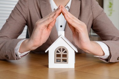 Photo of Property insurance. Real estate agent protecting house figure at wooden table indoors, closeup