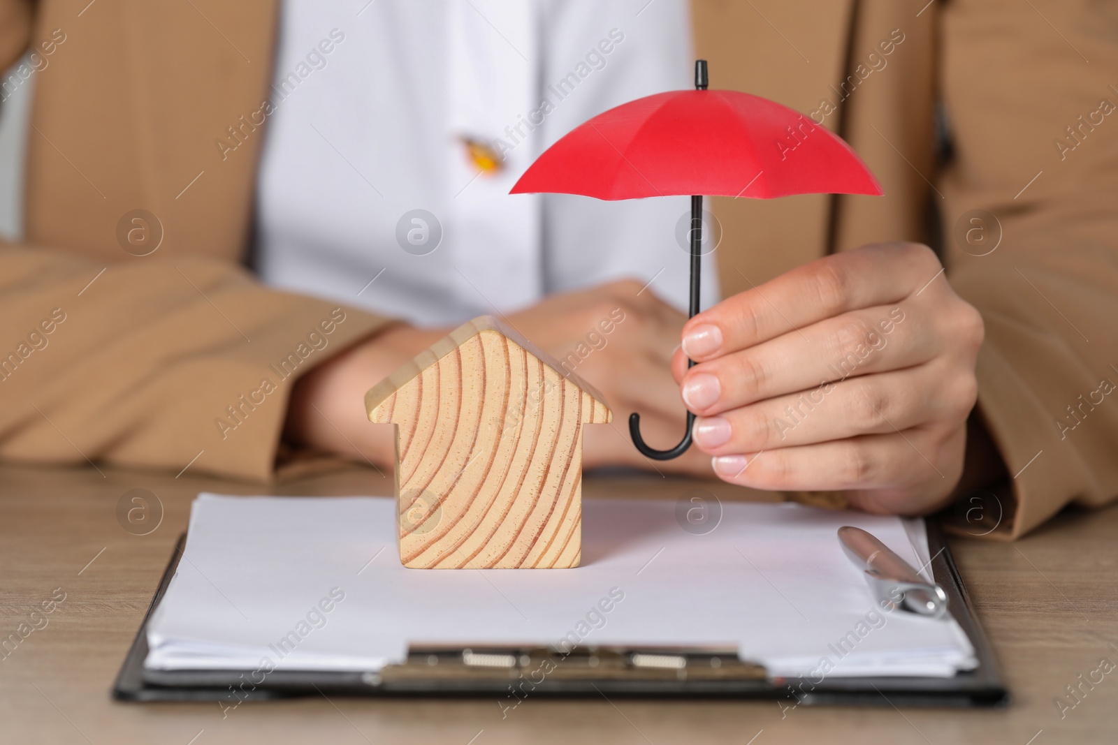 Photo of Property insurance. Real estate agent holding small umbrella above wooden house figure at table indoors, closeup