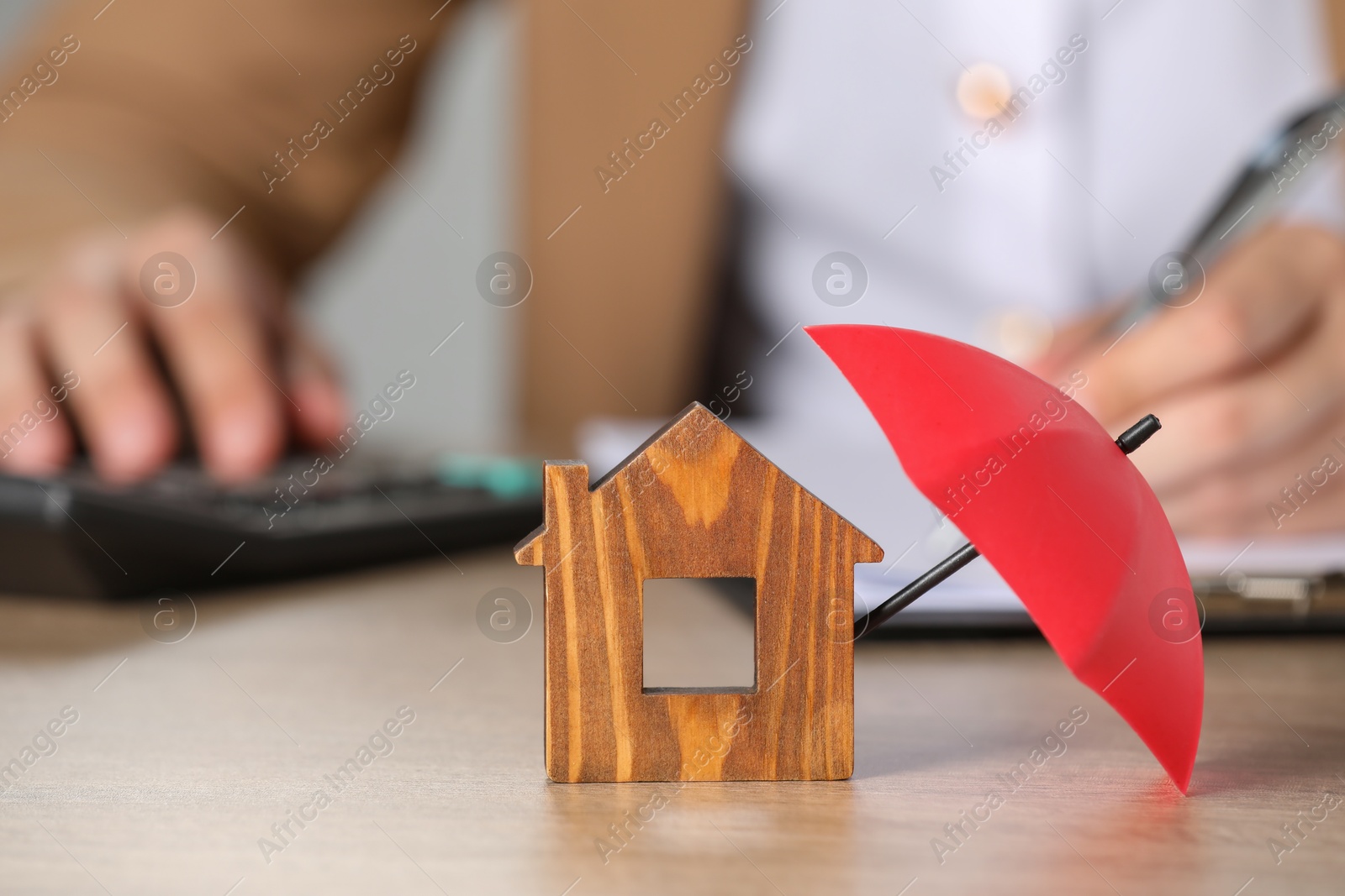 Photo of Property insurance. Real estate agent working at table indoors, focus on wooden house figure and small umbrella