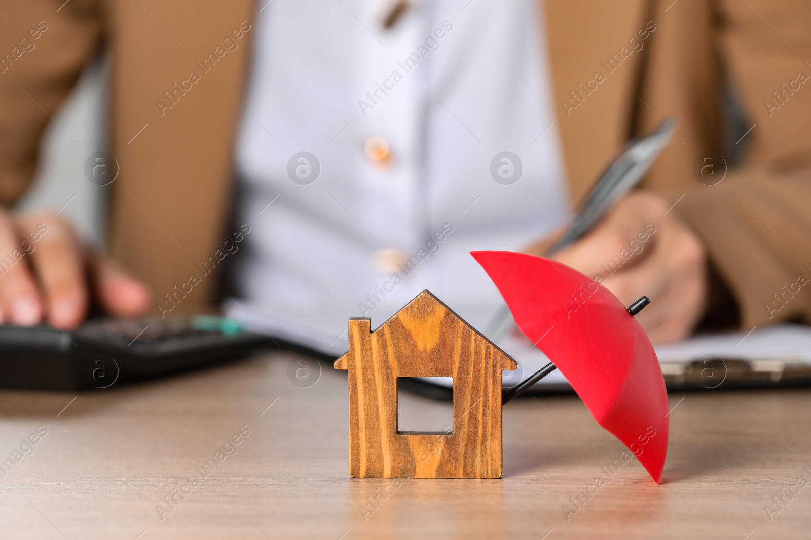 Photo of Property insurance. Real estate agent working at table indoors, focus on wooden house figure and small umbrella