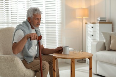 Lonely senior man with walking cane and cup of drink sitting at table indoors