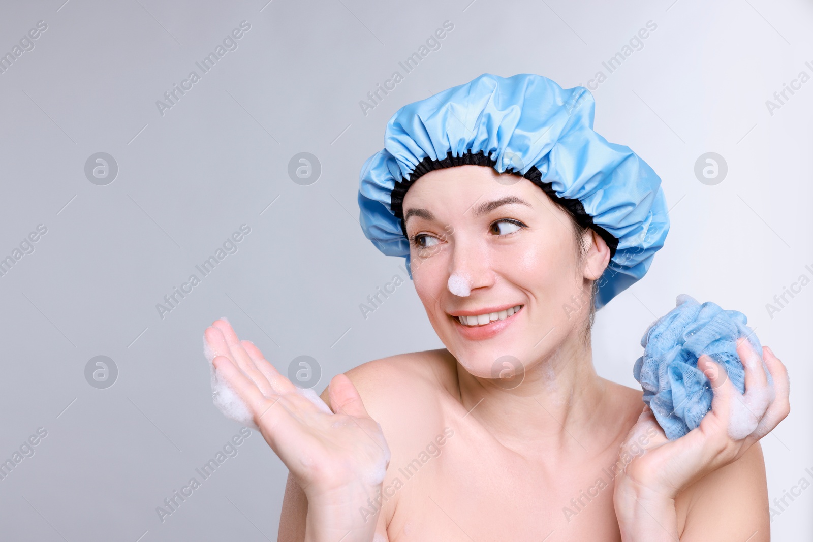 Photo of Woman in shower cap covered with soap foam holding mesh sponge on grey background