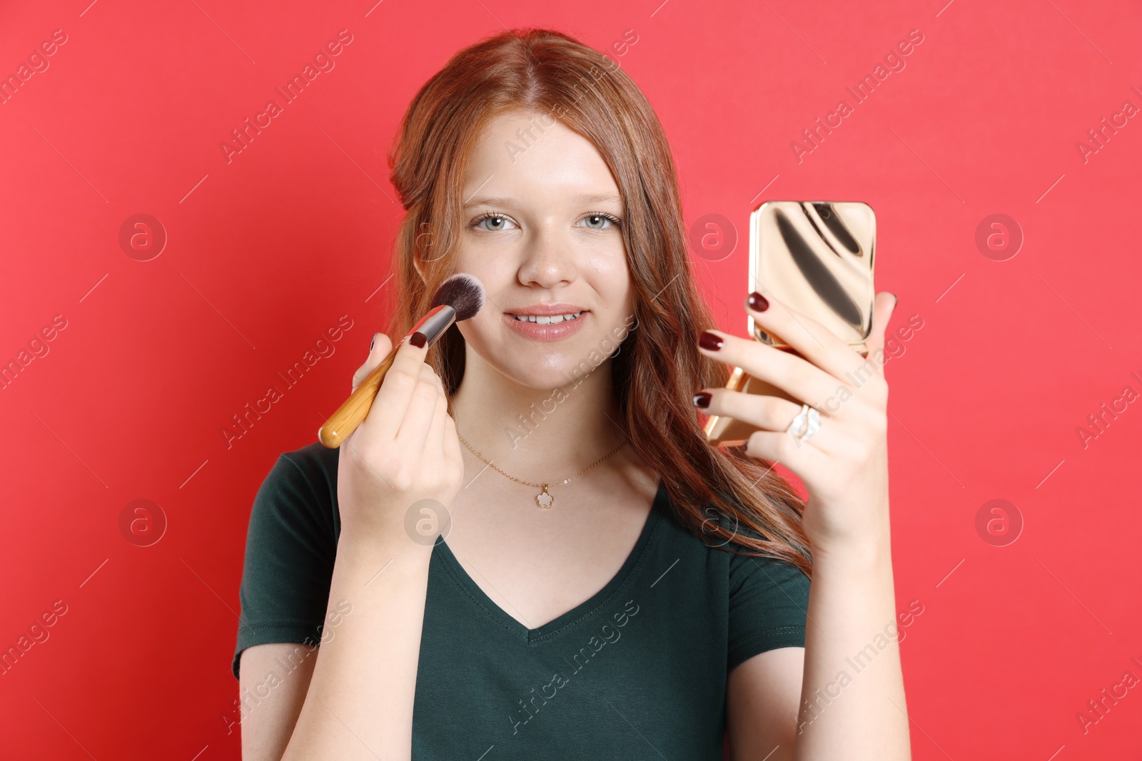 Photo of Smiling teenage girl applying blusher with makeup brush on red background