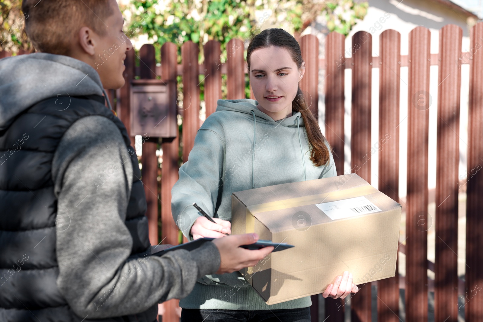 Photo of Woman signing papers while receiving parcel from postman outdoors