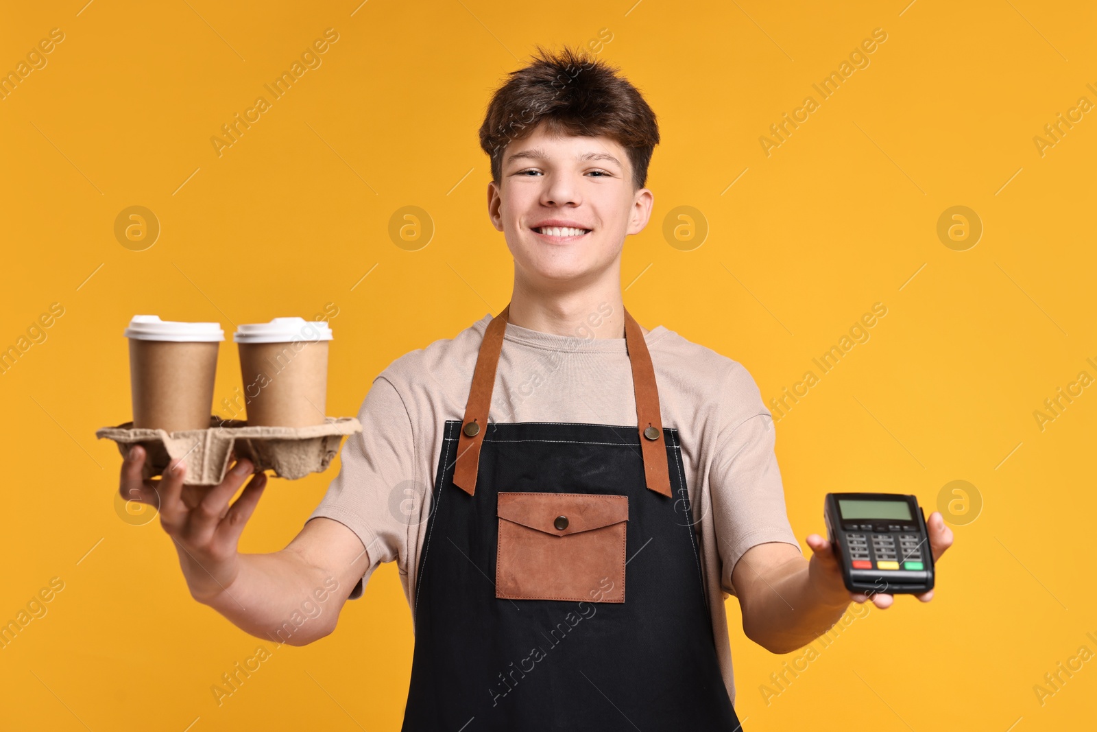 Photo of Teenage boy with paper cups and payment terminal working as barista on orange background