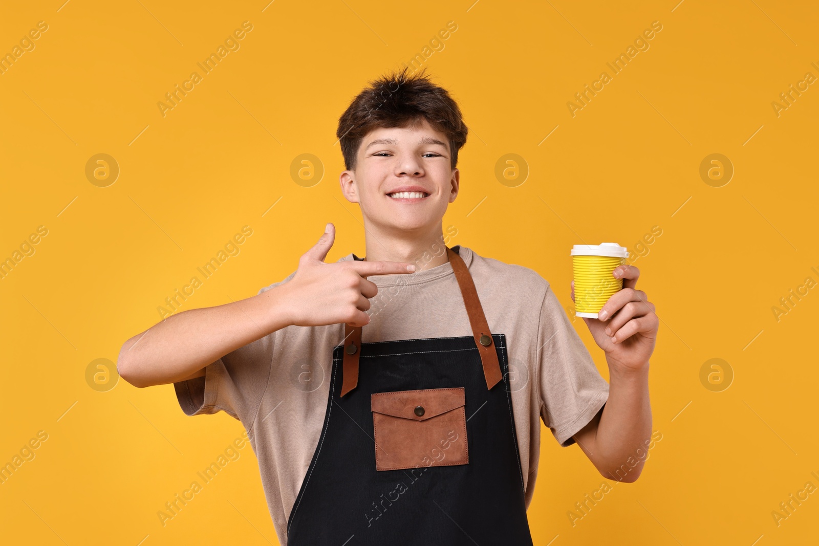 Photo of Teenage boy with cup of coffee working as barista on orange background