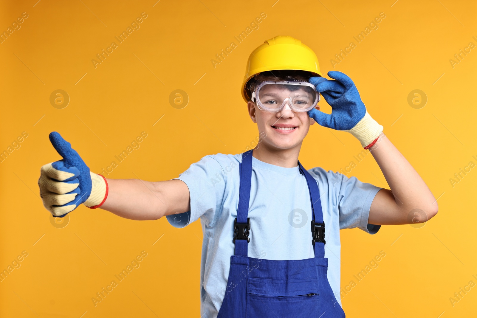 Photo of Teenage boy in hardhat and protective mask working as builder on orange background