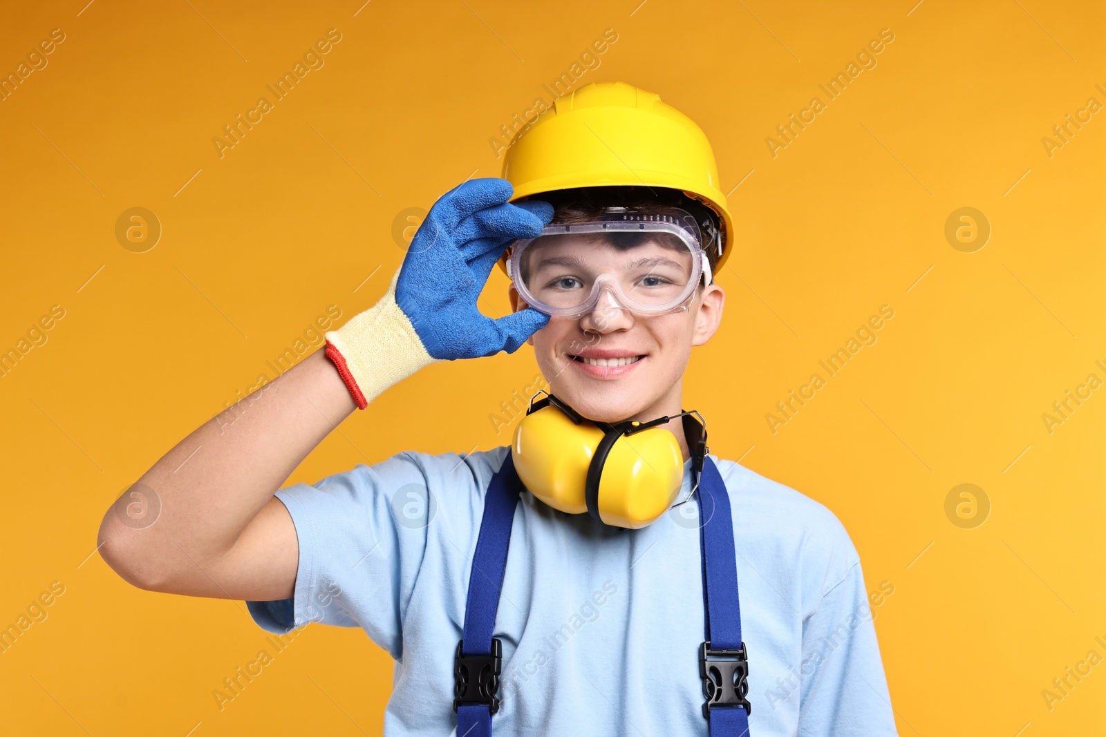 Photo of Teenage boy in hardhat and protective mask working as builder on orange background