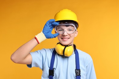 Photo of Teenage boy in hardhat and protective mask working as builder on orange background