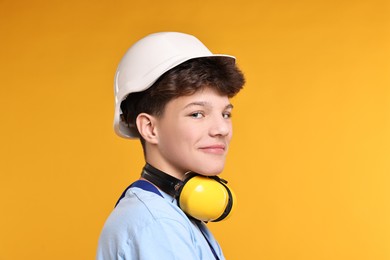 Photo of Teenage boy in hardhat with earmuffs working as builder on orange background