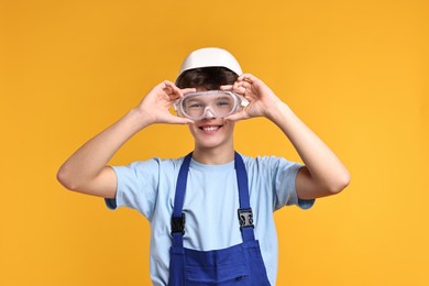 Photo of Teenage boy in hardhat and protective mask working as builder on orange background