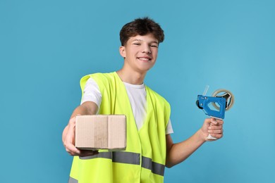 Photo of Teenage boy with tape gun dispenser and box in safety vest working as warehouse packer on blue background