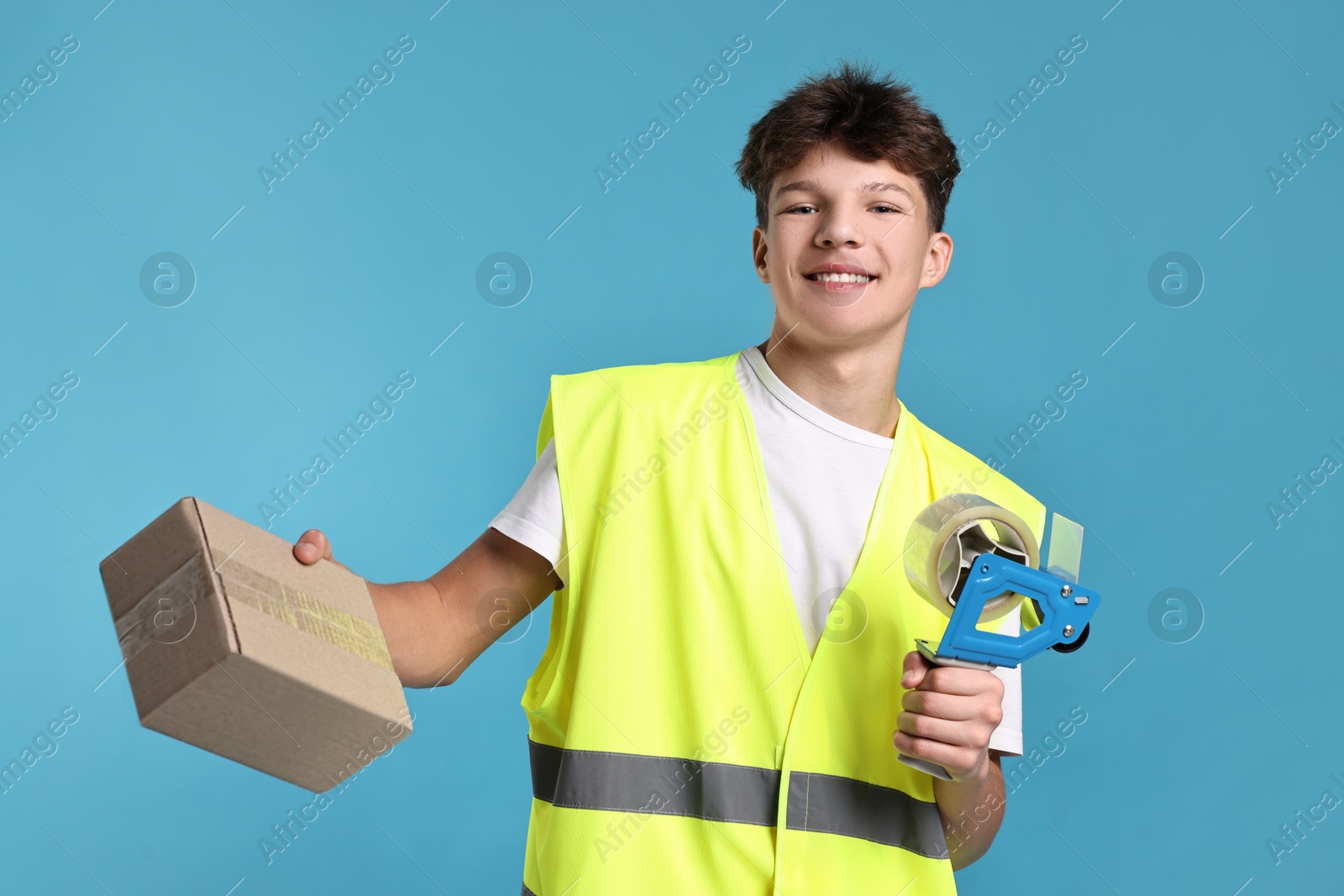 Photo of Teenage boy with tape gun dispenser and box in safety vest working as warehouse packer on blue background