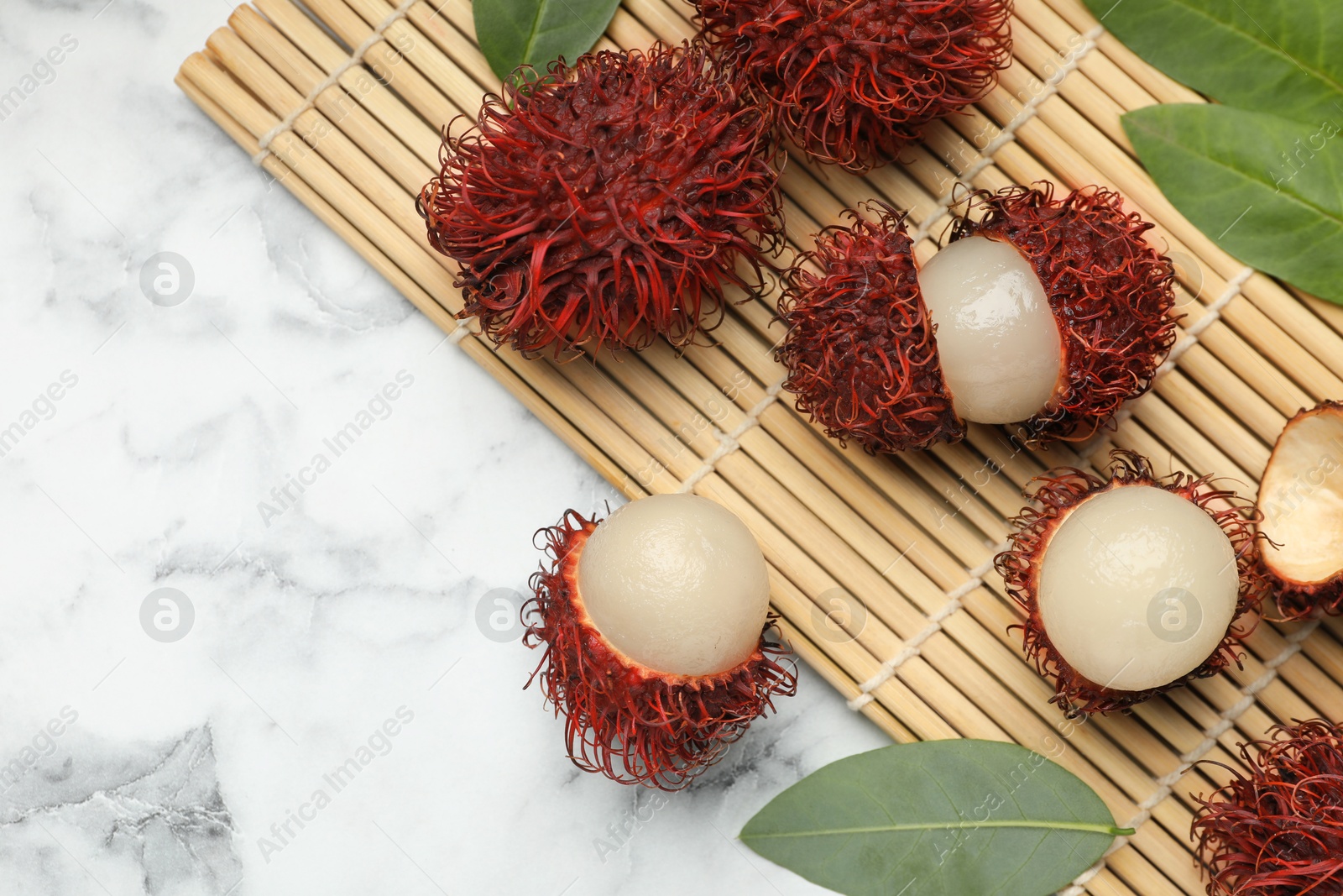 Photo of Delicious ripe rambutans and green leaves on white marble table, flat lay. Space for text