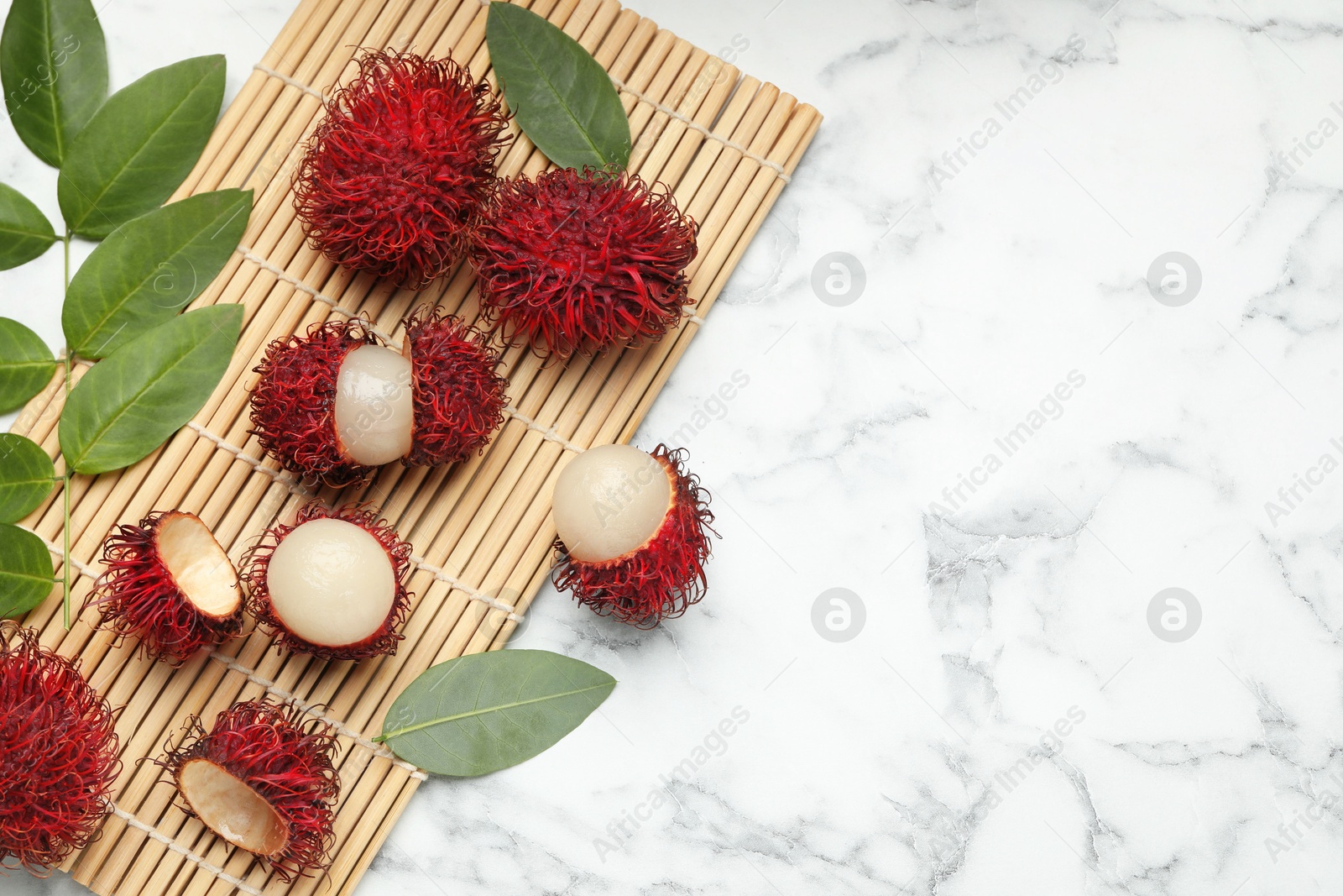 Photo of Delicious ripe rambutans and green leaves on white marble table, flat lay. Space for text