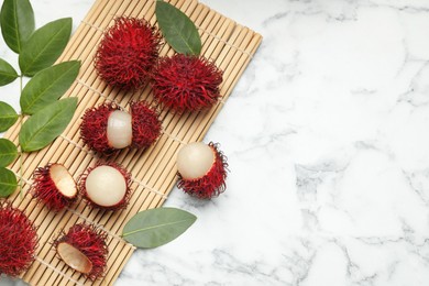 Photo of Delicious ripe rambutans and green leaves on white marble table, flat lay. Space for text