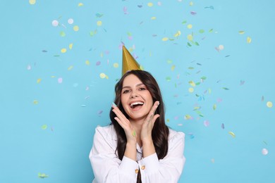 Photo of Happy woman in conical paper hat under falling confetti on light blue background. Surprise party