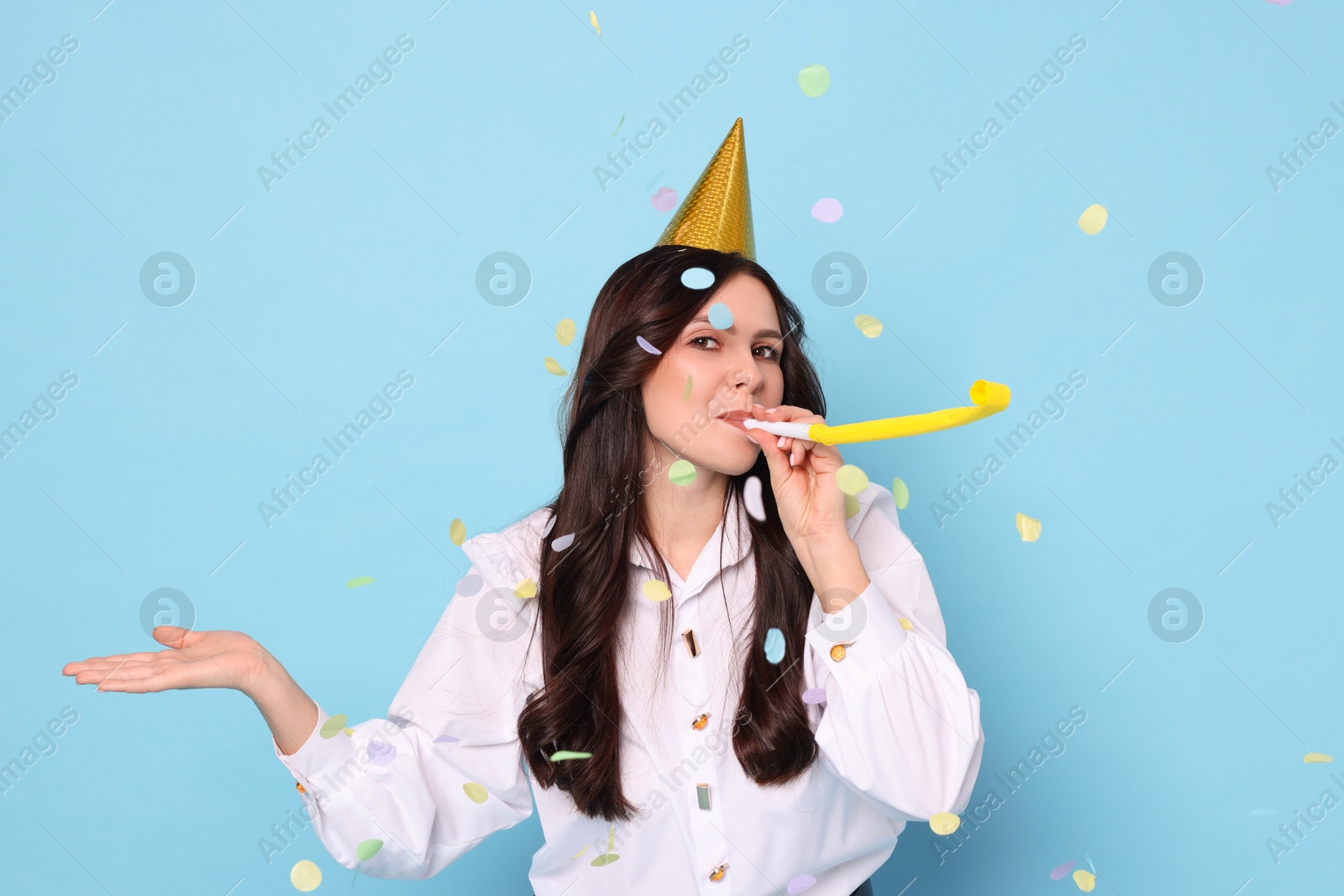 Photo of Woman in conical paper hat with blower and flying confetti on light blue background. Surprise party