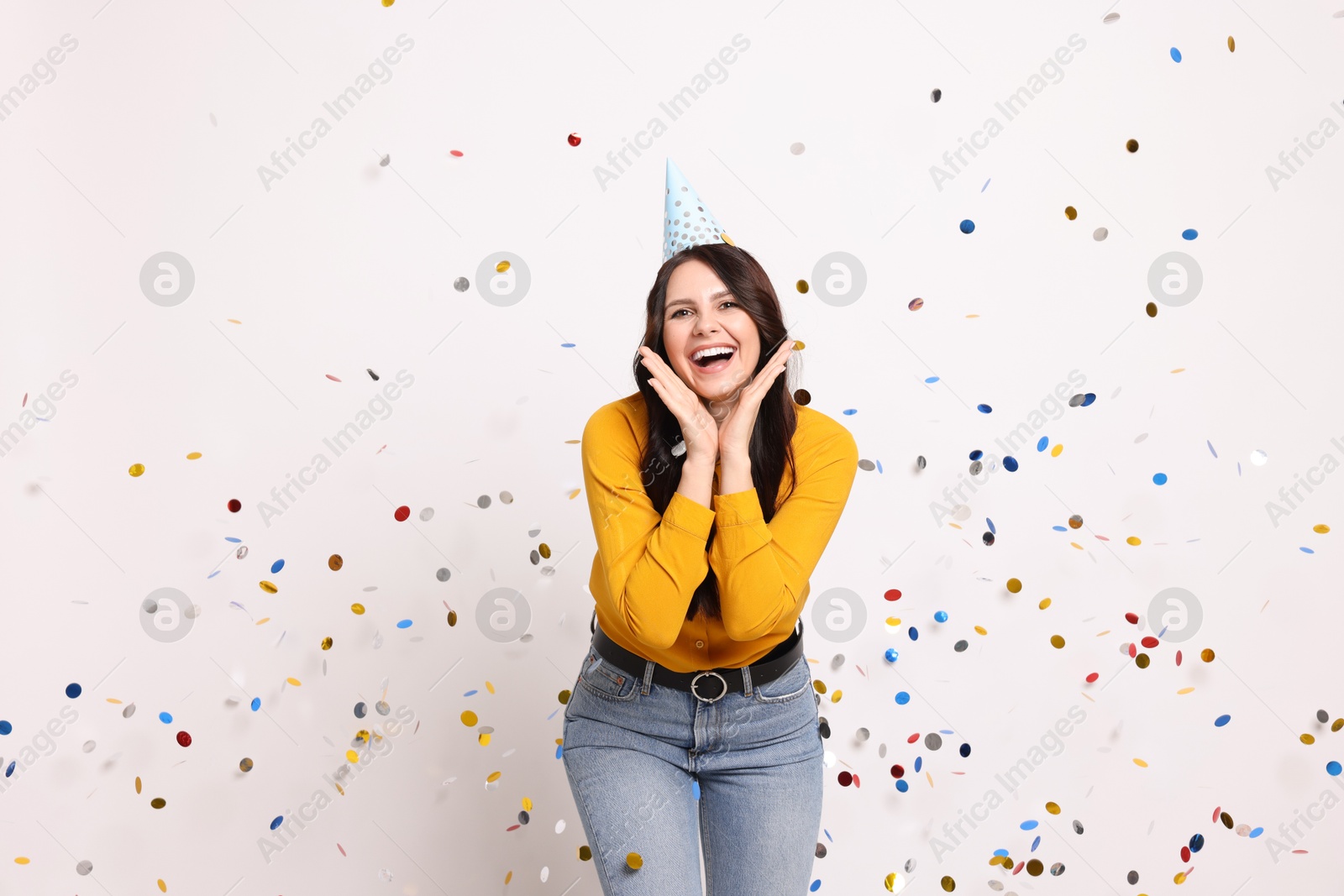 Photo of Happy woman in conical paper hat under falling confetti on white background. Surprise party