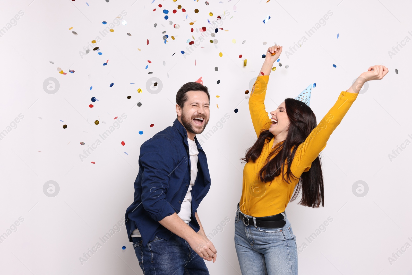 Photo of Happy friends in conical paper hats having fun under falling confetti on white background. Surprise party