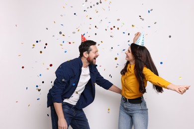 Photo of Happy friends in conical paper hats having fun under falling confetti on white background. Surprise party